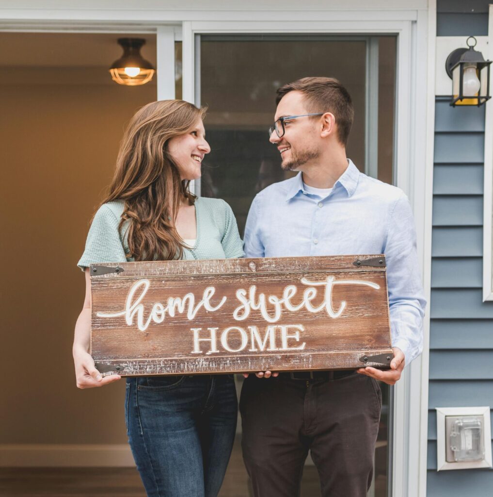 Young couple standing outside new home, holding 'home sweet home' sign, joyful and smiling.